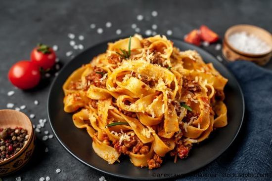 Nudeln mit Fleischsoße und Käse auf einem schwarzen Teller, Tomaten und Gewürze auf einer dunklen Oberfläche.