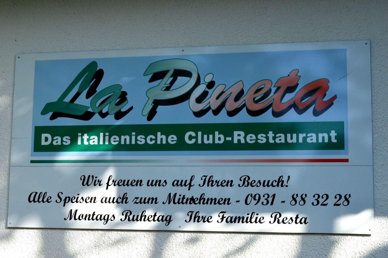 Schild für das italienische Restaurant „La Pineta“. Grün, Blau und Weiß. Mit Text zum Mitnehmen, Telefonnummer und Öffnungszeiten.
