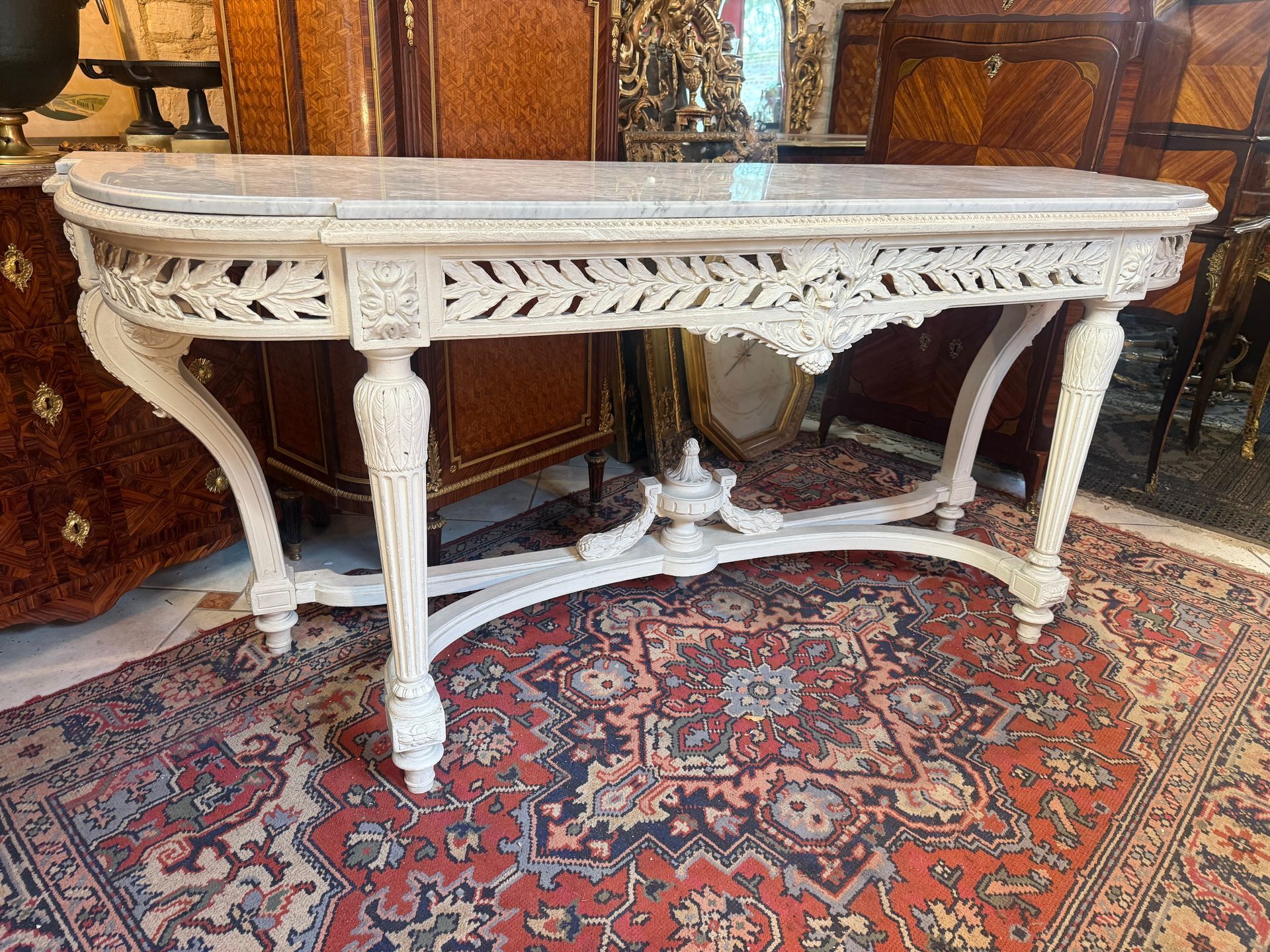Grande console d’apparat de style Louis XVI , En bois laqué patiné blanc avec dessus marbre de carrare. La ceinture richement ajourée figurant une large frise feuillagée. Le tout reposant sur des pieds cannelés, rudentés et ornés réunis par une entretoise surmontée d’une cassolette au centre sommé d’une pomme de pin. Provenant d’un appartement parisien haussmannien