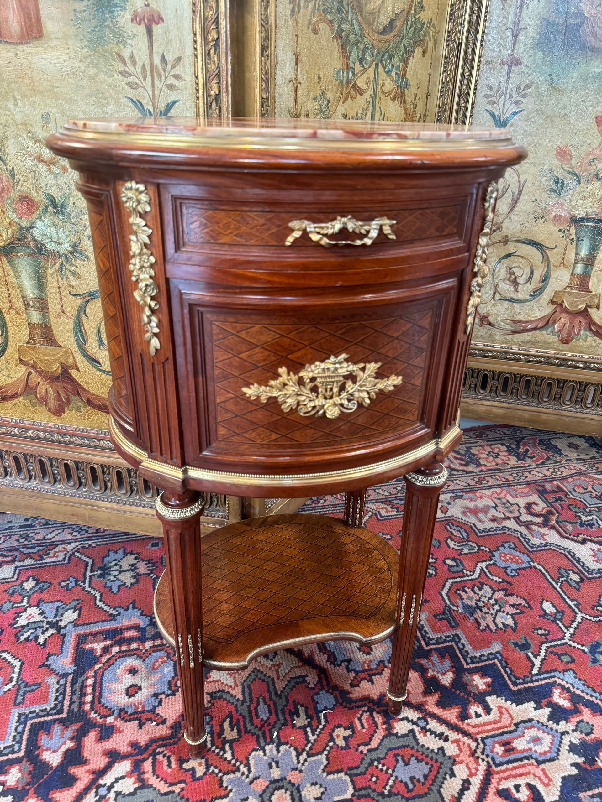 Table de chevet de forme tambour en placage d'acajou, de palissande et  marqueterie de losange, de style Louis XVI,
Cette table offre une belle ornementation de bronzes ciselés et dorés,
Elle ouvre par un tiroir et une porte et possède une tablette dans le bas,
le tout reposant des pieds cannelés,
Dessus marbre.
Signé F. Linke sur un bronze à la serrure en façade.
Meuble de grande qualité 
biographie : François Linke, né en Bohême (Tchécoslovaquie) en 1855, débuta comme ébéniste vers 1882 et  exerça à Paris jusqu’à sa mort, en 1946, au faubourg Saint-Antoine. Vers 1900, à l’apogée de sa carrière, il adjoignit même une succursale, place Vendôme. Il s’était spécialisé dans la fabrication de meubles de style Louis XV et Louis XVI, ambitieux tant par leurs dimensions que par leur somptueuse ornementation de bronzes, ce qui lui valut de nombreuses commandes dès la fin du XIXème s. Désirant aller au-delà des copies de style XVIIIème, Linke collabora avec le déjà célèbre sculpteur Léon Message et intégra les lignes sinueuses annonçant l’Art Nouveau, développant ainsi un style très personnel.
L’un de ses grands succès fut remporté à l’Exposition Universelle de 1900, où le jury lui décerna la médaille d’or pour son bureau, dessiné par Message, en bois de violette, monté de bronzes d’esprit Louis XV. La « Revue artistique et industrielle » glorifia Linke en écrivant que son stand à l’Exposition était la plus grande démonstration jamais réalisée dans l’histoire du mobilier d’art.