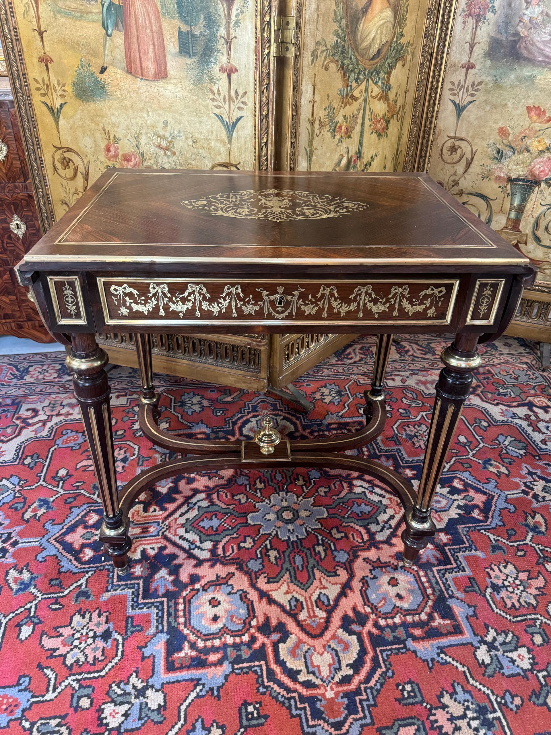 Jolie table à deux abattants aux extrémités, de style Louis XVI, époque Napoléon III, 19ème,
Table en placage de palissandre,
Elle présente un riche décor d’incrustations de laiton finement ciselé sur le dessus, en ceinture et sur les abattants ( Guirlande de nœuds Louis XVI, feuillages et fleurs, arabesques, urne fleurie ...)
Elle ouvre par un tiroir en ceinture et repose sur quatre pieds tournés à cannelures agrémentés de filets de laiton reliés par une entretoise surmontée dans son centre d’une cassolette.