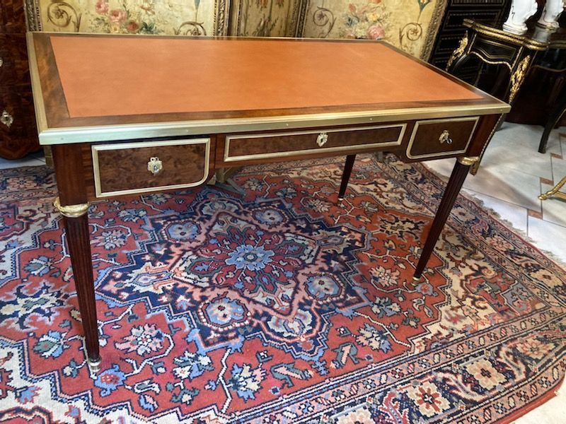 Elégant bureau plat toutes faces, de style Louis XVI - 19ème, 
En placage d'acajou et acajou moucheté, structure en chêne,
Ce bureau ouvre par trois tiroirs avec un décor d'une frise sur chacun des tiroirs et entrée de serrure avec nœud Louis XVI, 
Pourtour de frise sur chaque côté,
Il repose sur des pieds fuselés à cannelures, bague et sabots en laiton doré.
Le plateau est dessiné en ceinture par une lingotière  en laiton doré.
Le tout coiffé d'un cuir de couleur havane. 
Travail parisien de belle facture.