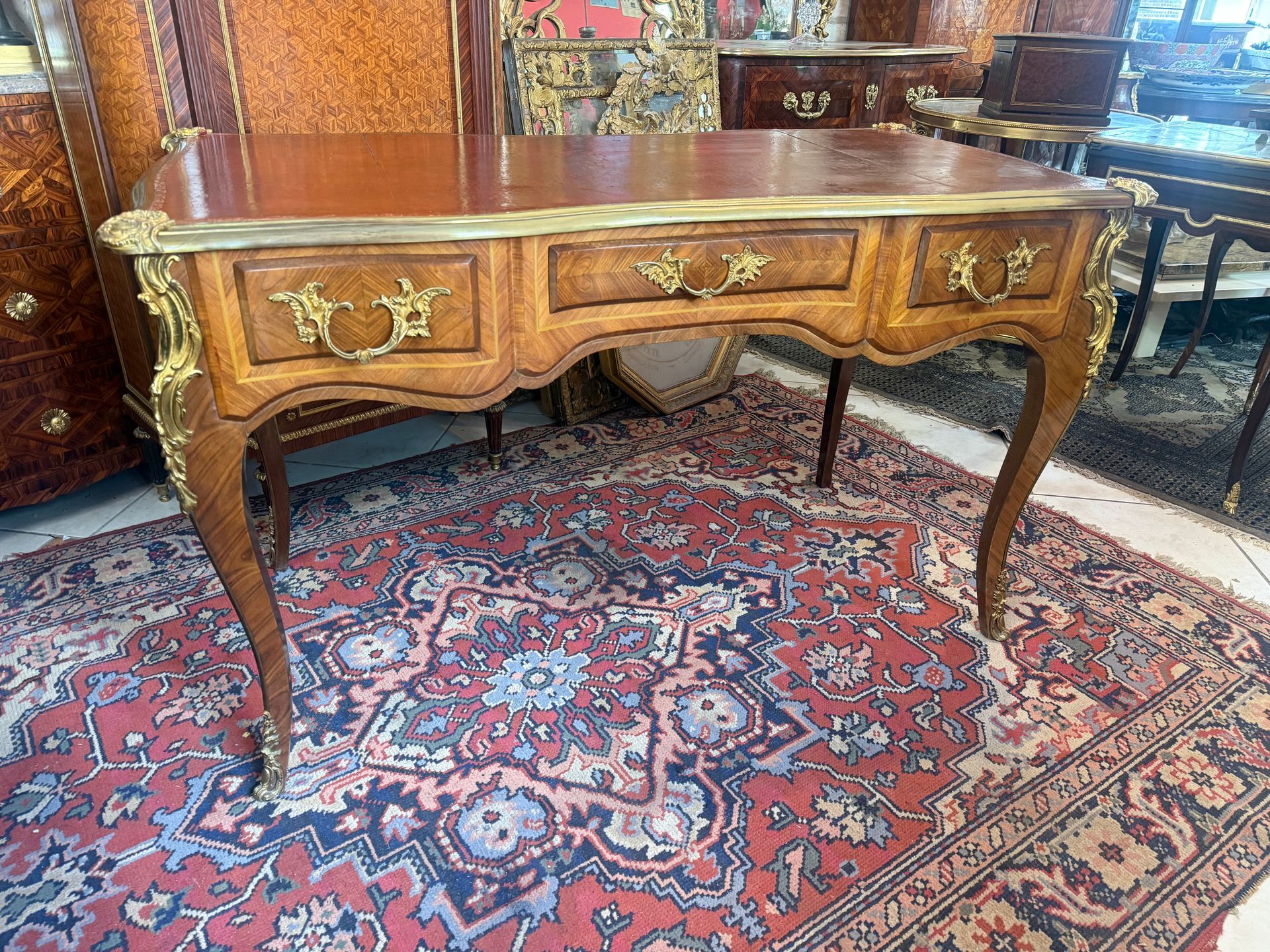 Bureau plat toutes faces de style Louis XV  En bois de placage de palissandre clair, Ouvrant à trois tiroirs avec poignées de tirage et tirettes de chaque côté, avec un encadrement de filets verts, Plateau rectangulaire chantourné, lingotière en laiton avec des mascarons de bronze à chaque angle,   Recouvert d’un cuir rouge d’origine, ainsi qu’un cuir de même couleur sur les tirettes. Le tout reposant sur des pieds cambrés avec décor de chutes de bronze doré sur les montants et les sabots. Travail parisien du faubourg St Antoine - début 20ème.