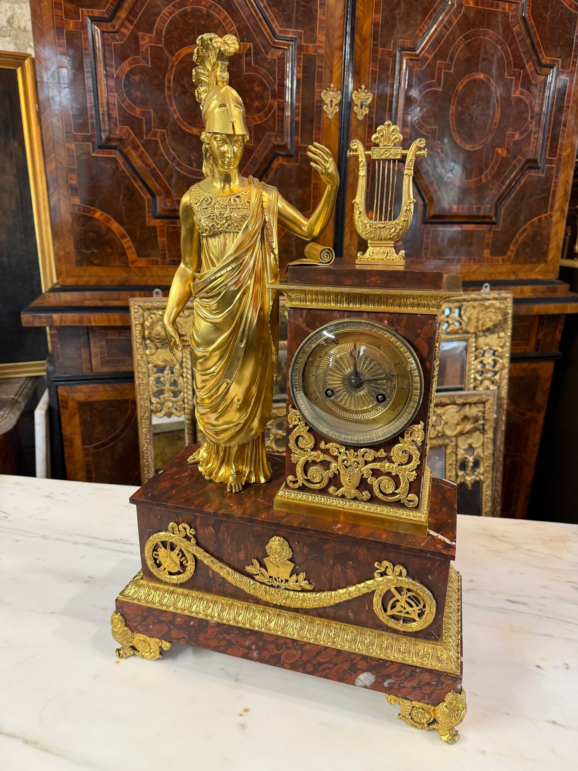 Grande pendule Empire début XIXème à décor d’une minerve casquée, de philosophe et de l’allégorie des arts.
Cette pendule est en marbre rouge et présente un riche décor de bronze doré et ciselé de belle qualité avec sa dorure d’origine .
 L’ensemble est supporté par une base de forme rectangulaire réunis par une couronne de laurier aux attributs des Arts, sur les côtés carquois de flèches,  pourtour en raie de cœur.
Elle repose sur des pieds en forme de chouettes.
Le mouvement est à fil.
