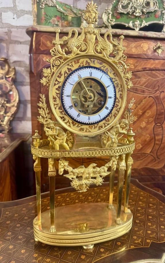 Pendule portique retour d’Egypte –Empire, En bronze doré,  Décor : Elle présente autour du cadran en partie haute au sommet un vase à l’antique avec  tête de méduse couronnée, Deux cygnes ailés et deux chérubins portant une branche de laurier, Décor floral ajouré, Pourtour d’une frise ajourée,  Elle repose en partie basse sur six colonnes aux bustes d’égyptiennes,  Dans son milieu un aigle royal aux ailes déployées,   Les colonnes reposent sur un socle de forme ovale entouré d’un pourtour d’une frise ciselée, Petits ronds. Cadran : Cadran porté par les chérubins, en émail blanc à chiffres romains, contour rond à motifs ajourés,   Possède son balancier avec undécor d’une tête de méduse.