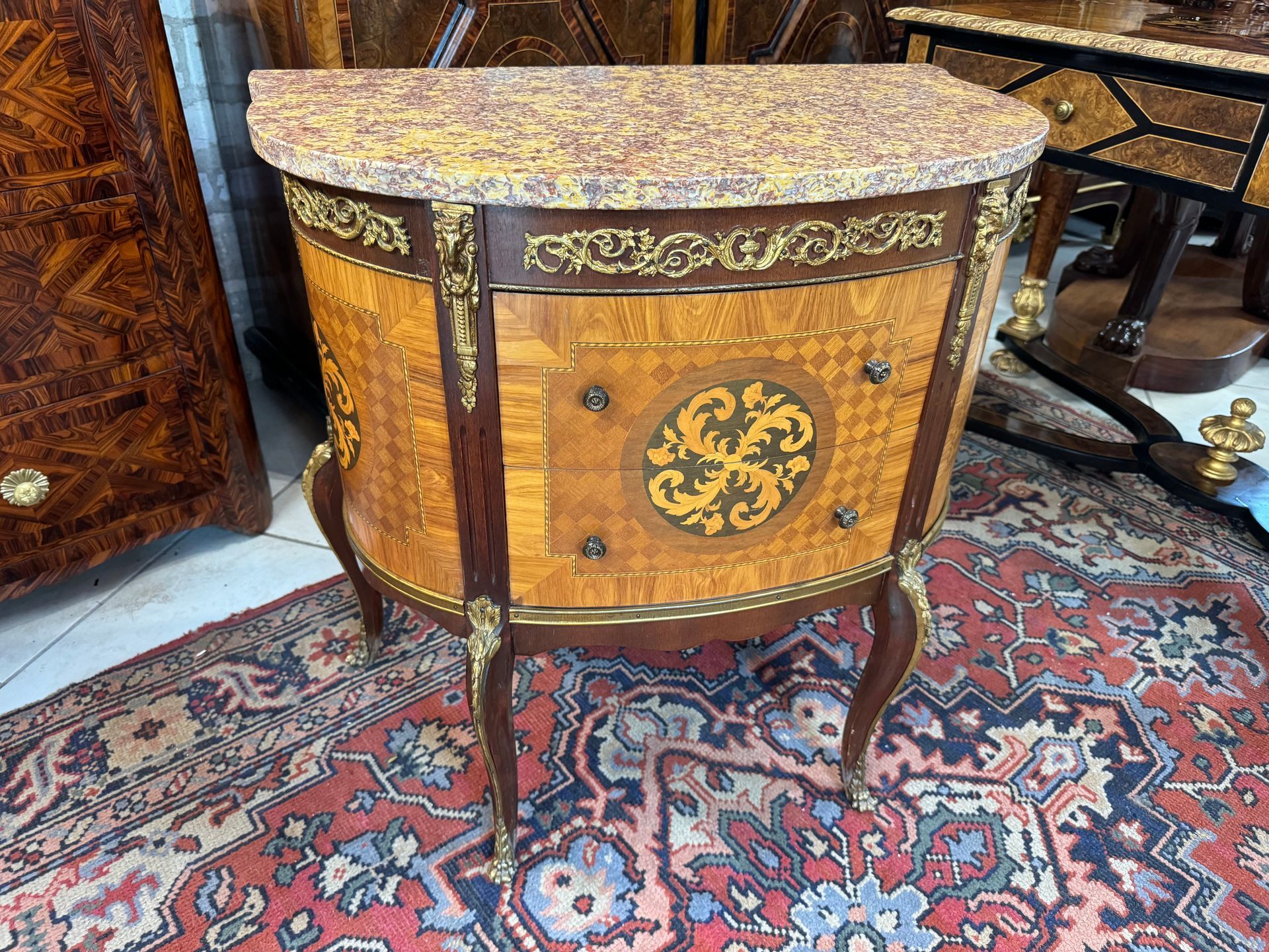 Elégante commode demi-lune de style LOUIS XV en marqueterie de bois de rose et palissandre.
Elle ouvre par deux tiroirs sans traverse ,  avec boutons petites fleurs.
Le devant et les côtés sont incrustés d'un décor de rosace,
Elle présente un décor de bronze formant tête de bélier sur les montants, frise de bronze en ceinture, bronze sur le montant des pieds.
Campant sur des pieds galbés et un dessus coiffé d’un marbre brocatelle violette.