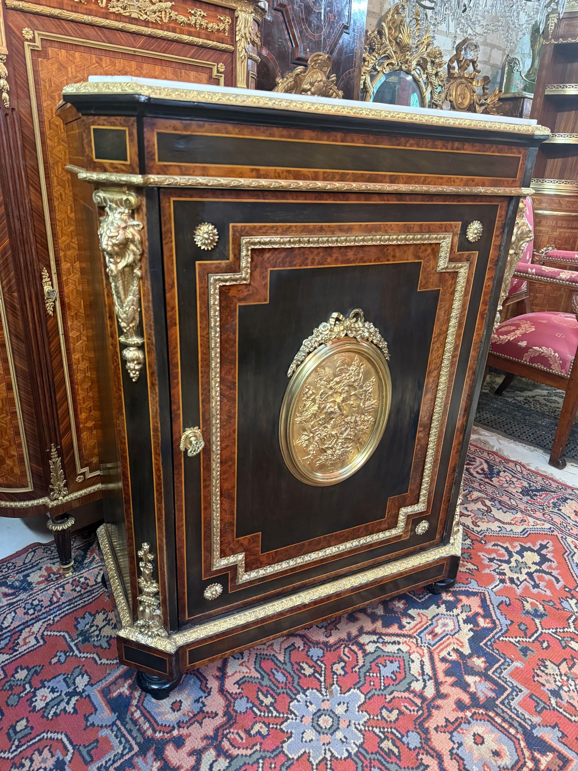Meuble à hauteur d'appui de style Louis XVl - Epoque Napoléon lll  ouvrant par une porte offrant deux étagères. En bois noirci, placage de loupe de noyer et fillets de buis, Décor d'un médaillon central en bronze repoussé représentant un couple de colombes, carquois, instrument de musique et panier de fleurs, Chute de tête de lion en bronze doré ciselé sur les montants,  Le tout coiffé d'un marbre blanc de carrare, entouré d'une lingotiere en bronze  Et reposant sur des pieds toupie. Meuble de belle qualité.