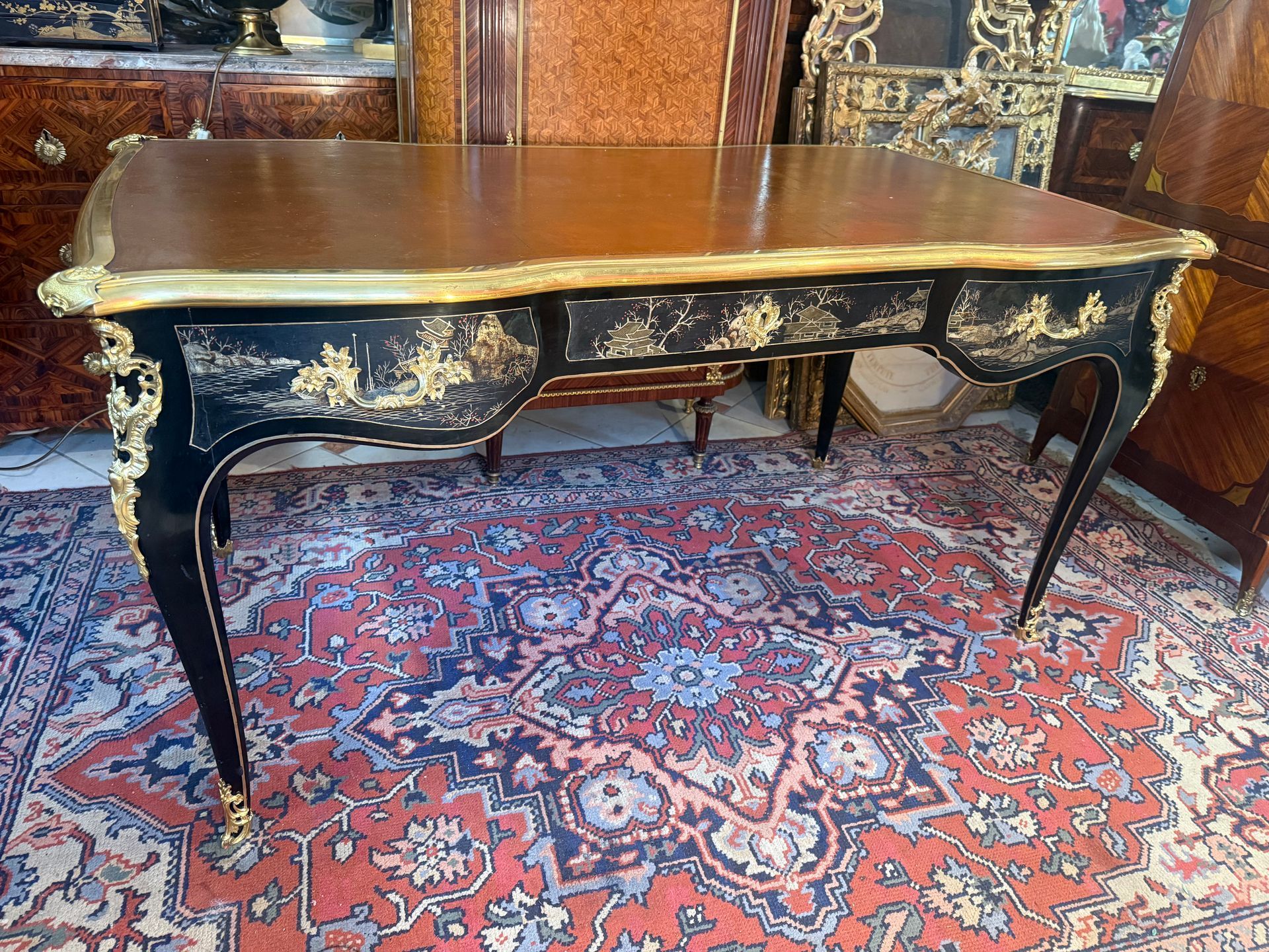 Bureau plat toutes faces de style Louis XV époque 19ème de forme mouvementée, en bois laqué noir, à décor de motifs japonisants. Dessus recouvert d’un cuir havane d’origine avec un pourtour d’ une large lingotière en bronze offrant à chaque angle un mascaron, Il repose sur des pieds cambrés avec chute de bronze finement ciselé sur les montants et les sabots. Dorure d'origine. Ce bureau ouvre à trois tiroirs soit deux petits de chaque côté avec poignée de tirage et un grand tiroir dans le milieu avec une entrée de serrure. Ce bureau japonisant est caractéristique de son époque et des créations de Perret et Vibert, les arts décoratifs de la fin du XIXe siècle témoignant d’un attrait renouvelé pour les créations extrême-orientales et s’amusant à détourner les codes de l’art japonais pour les appliquer à des meubles parfaitement occidentaux. A propos de : Alfred Perret fonde en 1879 àParis une maison spécialisée dans la réalisation de meubles en bambous etvannerie de luxe ainsi que dans l’importation d’objets authentiquement japonaisà destination d’une clientèle aisée et parisienne. La maison spécialisée dansla fabrication de meubles en bois sculpté d’inspiration extrême-orientale etmeubles en laque, prend le nom de « Maison des Bambous au début des années1890. 1884 : Le début des meubles japonisant A partir de 1884, l’activité se développe encore en s’étendant aux « meubles de fantaisie » et un an après est utilisée dans cette même catégorie la dénomination de « petits meubles de salon enbambou et laque, meubles chinois et japonais ». Nous situons ainsi à cette époque le début de la fabrication de meubles en bois sculpté, notamment pourceux d’inspiration asiatique. Certaines de ces créations seront d’un aspect formel très proche de Gabriel Viardot. Toutefois, la principale fabrication de la maison restera, jusque dans le milieu des années 1890 au moins, le fauteuil et la chaise en rotin et en bambou.