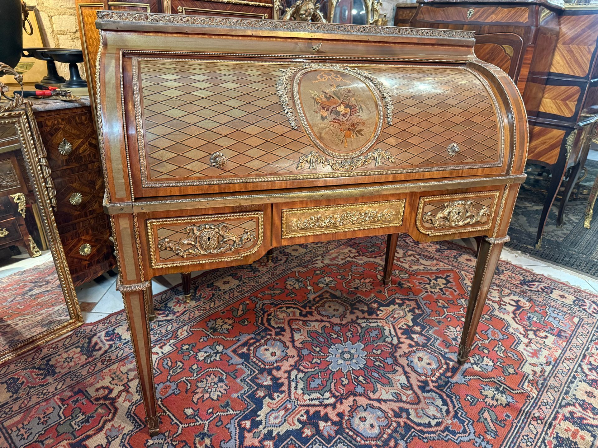 Bureau à cylindre de style Louis XVI d’après un modèle de Jean Henri RIESENER En marqueterie de losanges toute face, Le cylindre est orné au centre d’un médaillon représentant une allégorie sur le thème de la musique, entouré d’une guirlande de fleurs ciselée autour de ce médaillon et nœud Louis XVI Ce bureau offre un intérieur renfermant cinq tiroirs et trois niches ainsi qu'une tablette d’écriture se tirant recouverte d’un cuir havane, La ceinture ouvre à trois tiroirs dont un secret avec un bouton à poussoir, à riche ornementation de bronzes dorés à frises de putti et entrelacs feuillagés. Sur le dessus, galerie en laiton ajouré avec un plateau enmarbre de carrare blanc veiné, Repose sur des pieds gaine, Travail du Faubourg Saint Antoine début 20ème Le modèle de ce meuble est le bureau du Roi Louis XVI réalisé par Riesener et conservé aujourd'hui au Louvre.