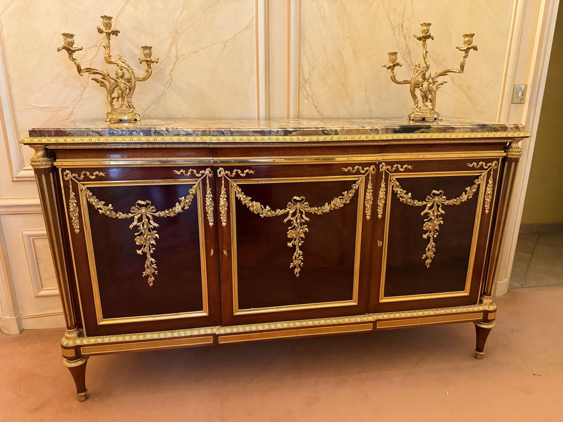 Buffet – Enfilade de style Louis XVI – 20ème siècle, en acajou massif, placage d’acajou et structure en chêne,
Offrant un riche décor de bronzes finement ciselés et dorés, formant des guirlandes de fleurs, nœuds Louis XVI, frises moulurées en façade ainsi que de frises sur chaque côté,
Façade droite ouvrant à trois vantaux
Reposant sur des pieds toupies à sabots et bagues de bronze, supportant chacun un montant en colonne cannelée.
Le tout coiffé d’un épais marbre brèche de Benou encastré dans un pourtour d’une frise en bronze doré.
Beau travail français, dans le goût de l’ébéniste Adam WEISWEILER