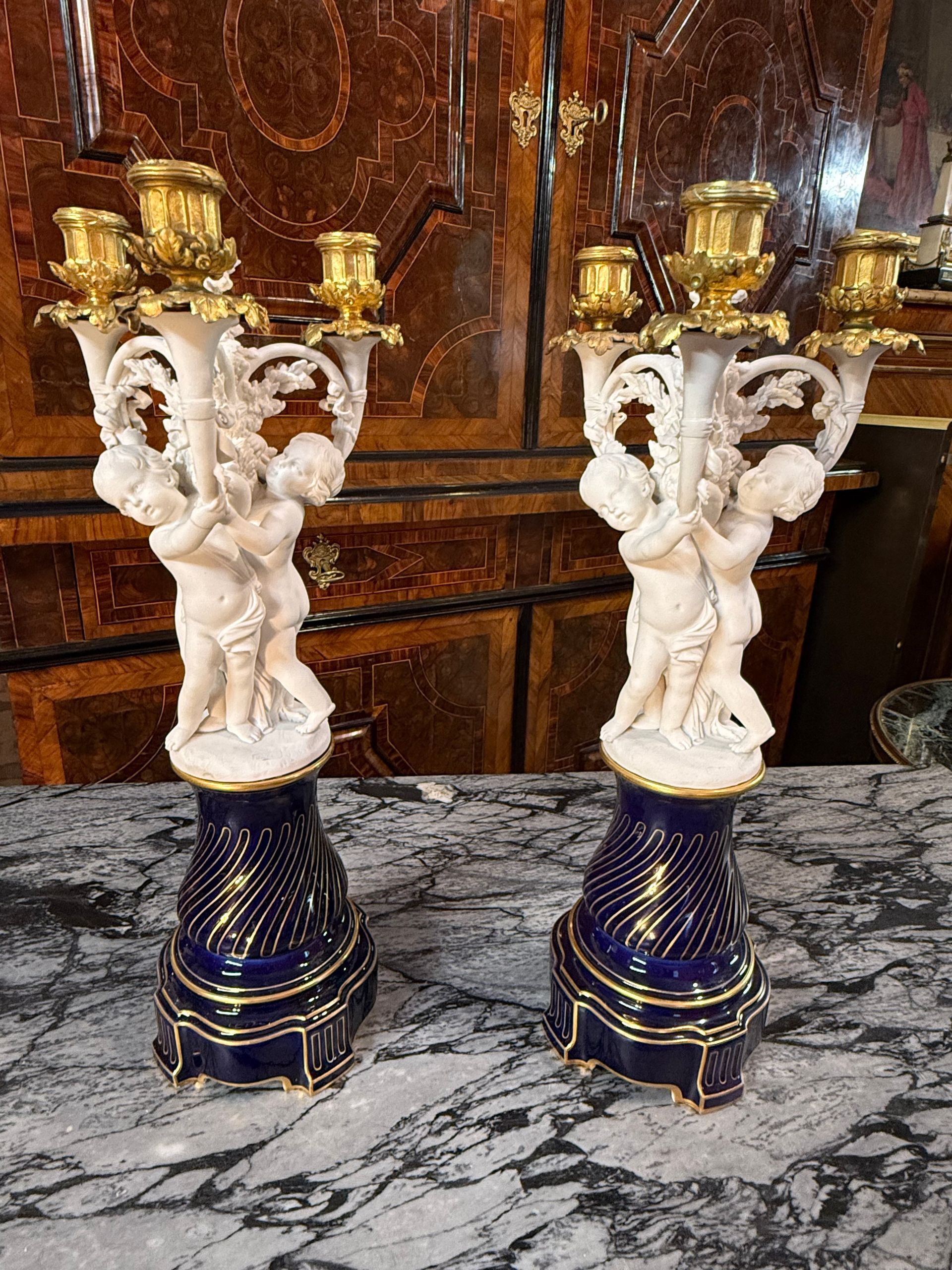 Jolie paire de candélabres en biscuit de sèvres, porcelaine et bronze doré, formés de putti soutenant des cors de chasse formant bras de lumière sur un socle bleu roi à godrons torses, bobèches formant un feuillage de chêne.
Sèvres, 1887
Numéroté et signé SEVRES 
Candélabres de qualité, décoratifs et de belle facture.