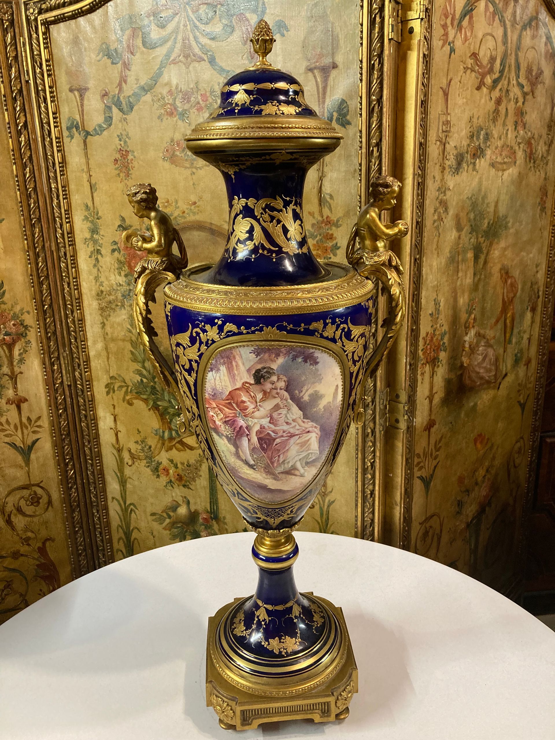 grand vase avec son couvercle en porcelaine de Sèvres 
Ce vase est de forme ovoïde à col évasé, surmonté de son couvercle lequel se termine par une pomme de pin et reposant sur socle en bronze doré avec quatre petits pieds toupie,
Le décor finement peint représente :
- Une scène galante sur le devant et une scène de paysage au revers, sur fond bleu cobalt,
- De chaque côté des putti en bronze doré finement ciselé jouant des cymbales,
- Sur l’ensemble un travail à la dorure finement exécuté à décor de feuilles, fleurs stylisées et guirlande de perles.
Ce vase est très décoratif par son décor et sa grandeur.
