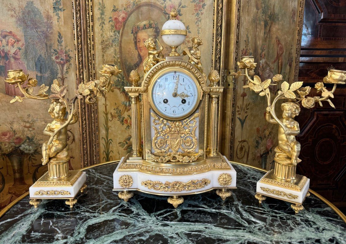 Garniture de cheminée,  de style Louis XVI époque Napoléon III,
Cette  pendule harmonieuse et romantique par son décor,  de forme borne présente un riche décor de bronze doré finement ciselé et exécuté,
On y voit ;  
-  socle en marbre de carrare,  supportant deux colonnes de chaque côtés se terminant par des pommes de pin,
-  le tout surmonté d'une boule sphérique réunie par deux angelots jouant de la musique,
 - décor de guirlandes de fleurs, perles,  mascarons et carquois  etc... dans son ensemble.
- cadran émaillé avec une guirlande de petites fleurs roses et feuillage mettant en valeur les chiffres, mouvement de Paris, 
Le tout accompagné par deux chandeliers  à deux bras de lumière, socle en marbre de carrare,  supportant  un angelot  assis sur un tronc portant en main une branche de roses. 
Envoi possible en Colisimo dans toute l'Europe.
Dimensions ;
La  pendule 43 de haut 28 de large et 15 de profondeur.
Les  chandeliers : 30 de haut et 9 de profondeur.
