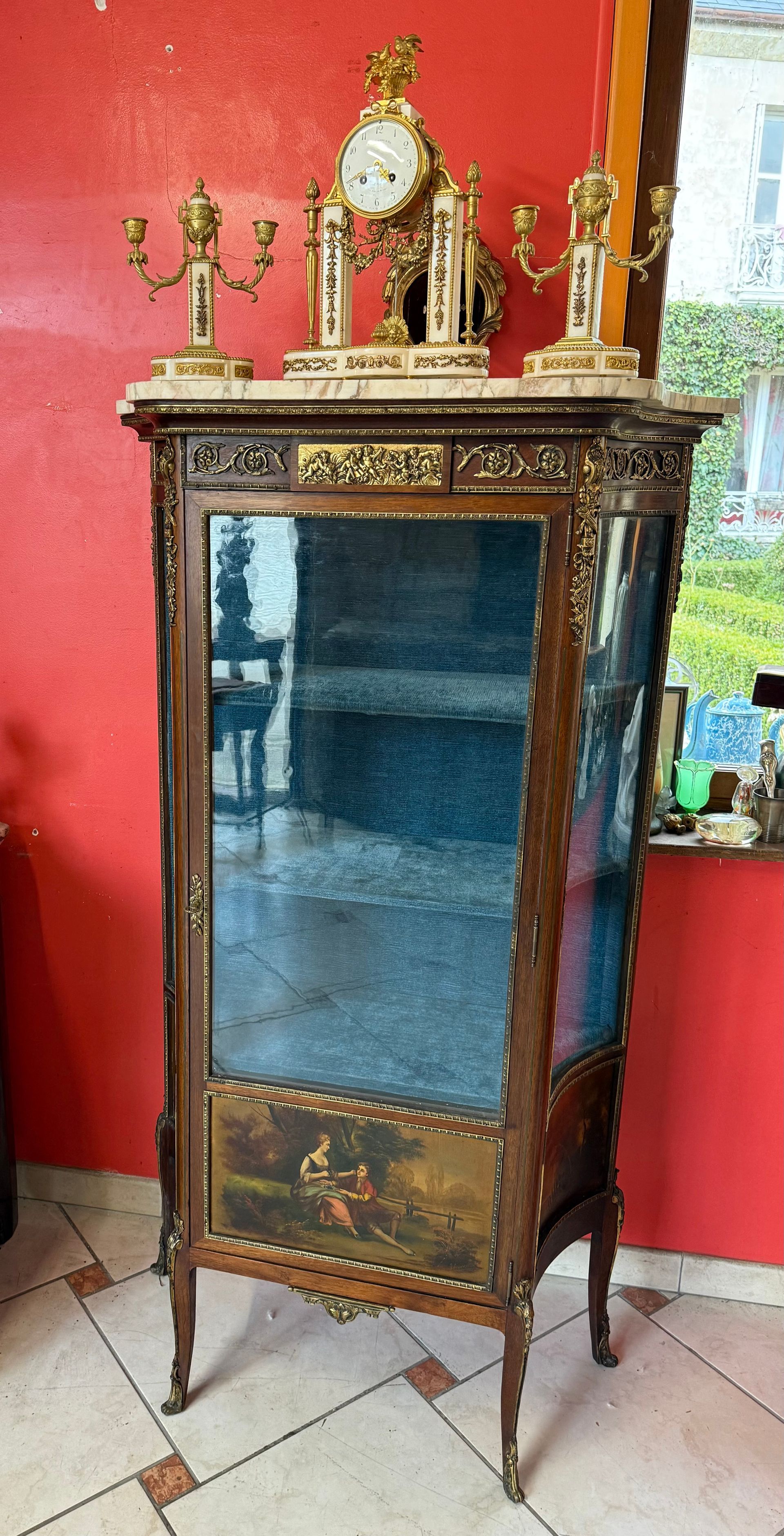 Vitrine galbée d'époque Napoléon III - 19ème, ouvrant à une porte et deux étagères en verres, orne d'un décor de bronzes dorés ciselés représentant une scène de putti, guirlande de fleurs, noeuds, carquois, pourtour de frises,  etc...  Elle offre dans la  partie basse d'une scène galante en vernis martin. Cotés incurvés , Reposant sur des pieds dit carbrioles. Le tout coiffé d'un marbre blanc veiné.