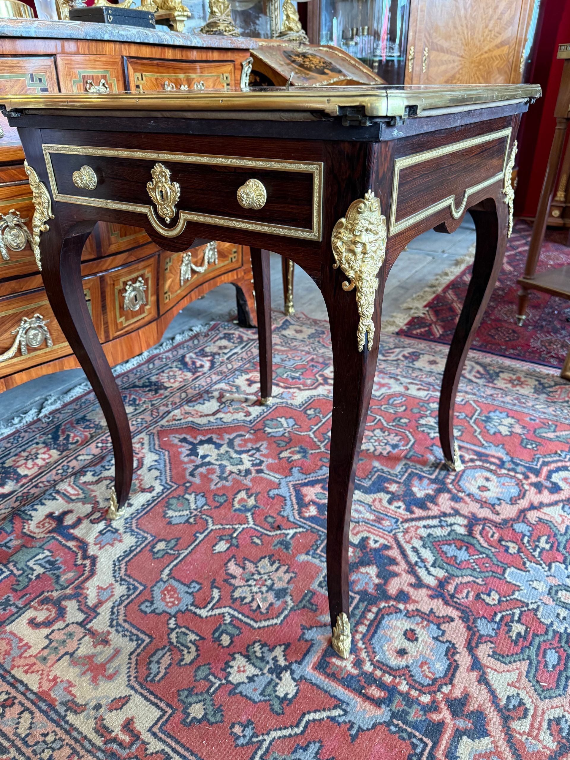 TABLE A JEUX EN MOUCHOIR EN PLACAGE DE PALISSANDRE 19EME ,  Elle offre un plateau    carré pivotant à quatre feuilles avec lingotière de bronze doré, souligné par    des baguettes, découvrant un tapis de jeu en velours vert   Elle ouvre à deux tiroirs    en ceinture avec deux petits boutons de tirage et entrée de serrure, entouré de    baguettes en bronze doré,  ainsi que des    baguettes de chaque côté. Elle repose sur des pieds galbés avec une ornementation de    bronze à décor de chute de tête d’homme barbu aux angles Estampillée Lampre Paris Travail parisien de belle facture. A propos de : A. LAMPRE, ébéniste spécialisé notamment    dans la fabrication de tables à jeux de tous styles pour lesquelles il détenait    un brevet. Il participa aux Expositions universelles de 1878 et 1889 où il    obtient plusieurs médailles de bronze. Il exerça au 72, faubourg Saint-Honoré à    partir de 1876 et 40, rue Amelot de 1882 à la fin du siècle