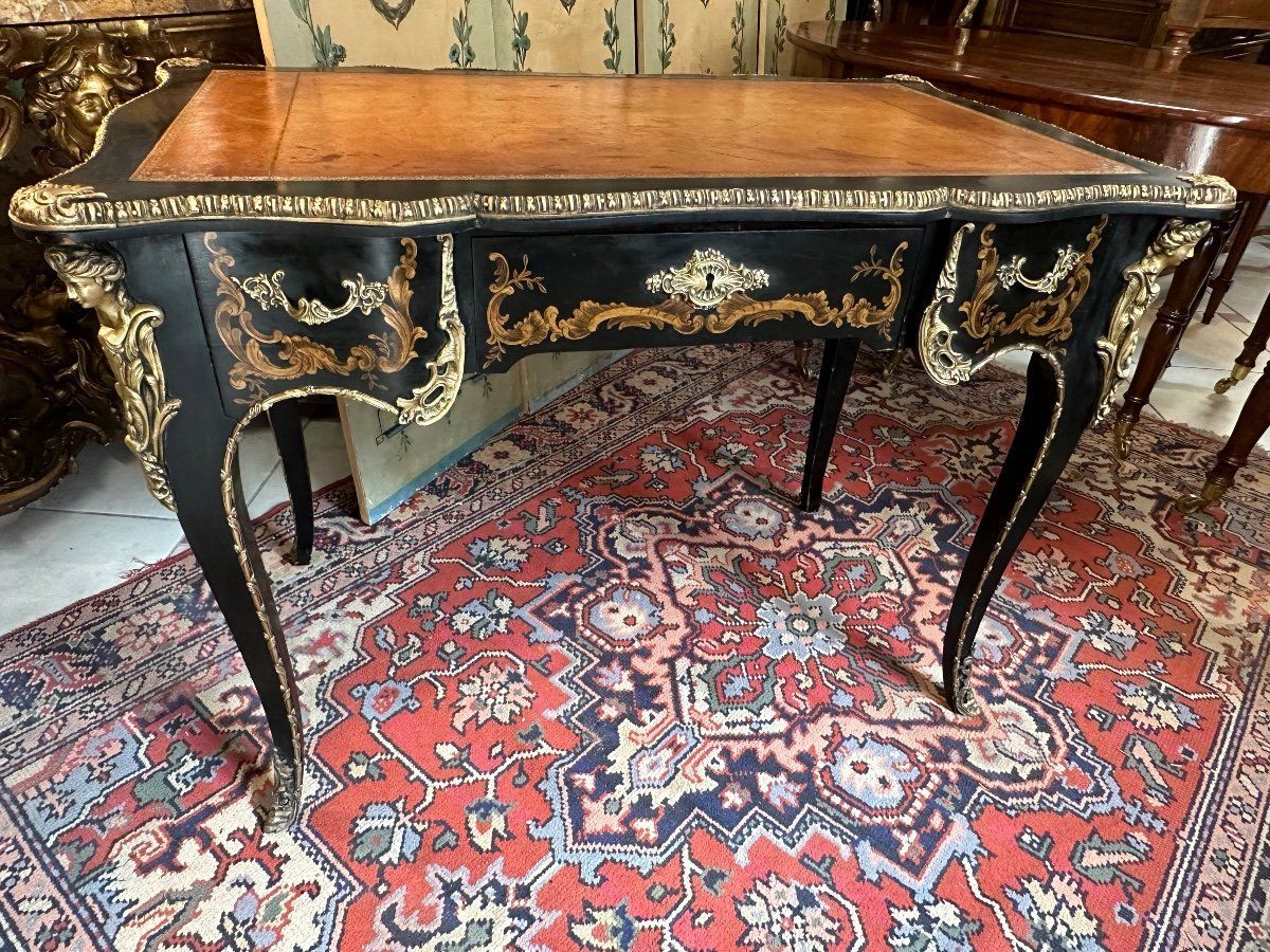 Bureau plat en bois laqué noir, de style Louis XV, avec un décor à la penture.
Cet élégant bureau est de forme légèrement mouvementé, toute face, ouvrant sur une des faces par trois tiroirs avec poignées de tirage.
Il présente dans son ensemble une riche ornementation de bronze doré.
Il repose sur quatre pieds cambrés avec de chûtes de tête de femme à l'antique.
Le plateau est encadré d'une lingotière en bronze mouluré et recouvert d'un cuir de couleur havane.