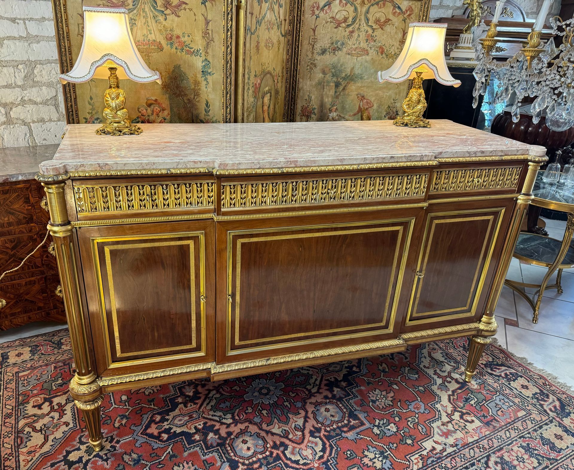 Elégant buffet - enfilade en acajou et placage d’acajou,
Structure chène.
Ce buffet ouvre par trois vantaux,  offrant un intérieur de rangement organisé soit une rangée de cinq tiroirs sur la gauche avec chacun un écrin pour aménagement d'argenterie dans son milieu et sur la droite, un rangement avec une étagère chacun.
Il est riche en ornementation de bronze finement ciselé a savoir, 
- en ceinture et sur les côtés,
-  sur les moulures de frise aux encadrements des portes, sous le marbre.
Ornementations de laiton sur les  montants en colonne cannelée et pieds.
Le tout reposant sur quatre pieds dit « toupie »
Le tout coiffé d’un marbre encastré brêche veiné.
Travail parisien de belle facture,
 D'après un modèle d’ Adam Weisweiler. Ebéniste parisien (1744-1820) démarqué par ses meubles repris d’après l’Antique, lequel s’est installé rue du faubourg Saint Antoine à PARIS.