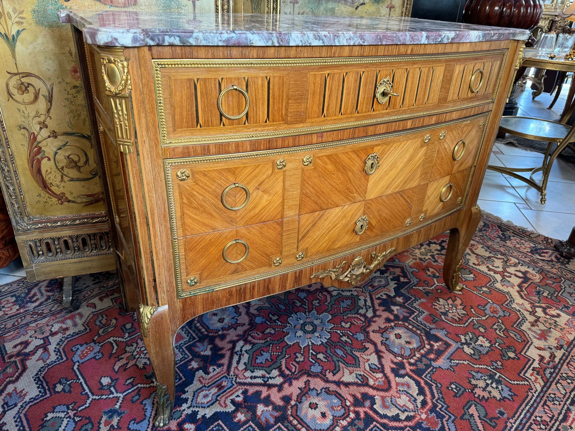 Belle commode style Transition Louis XVI en placage et bois de rose.
Elle présente un travail de marqueterie à décor symétrique,   une ornementation de bronze doré, frise de perles, petites rosaces, chute d’angle, cul de lampe à décor de cassolette
Elle ouvre en façade à trois tiroirs   sur trois rangs, anneaux de tirage à décor de perles, serrure médaillon de nœud Louis XVI.
 Elle repose sur quatre pieds cambrés à sabots.
Le tout coiffé d’un dessus de marbre fleur de pêcher