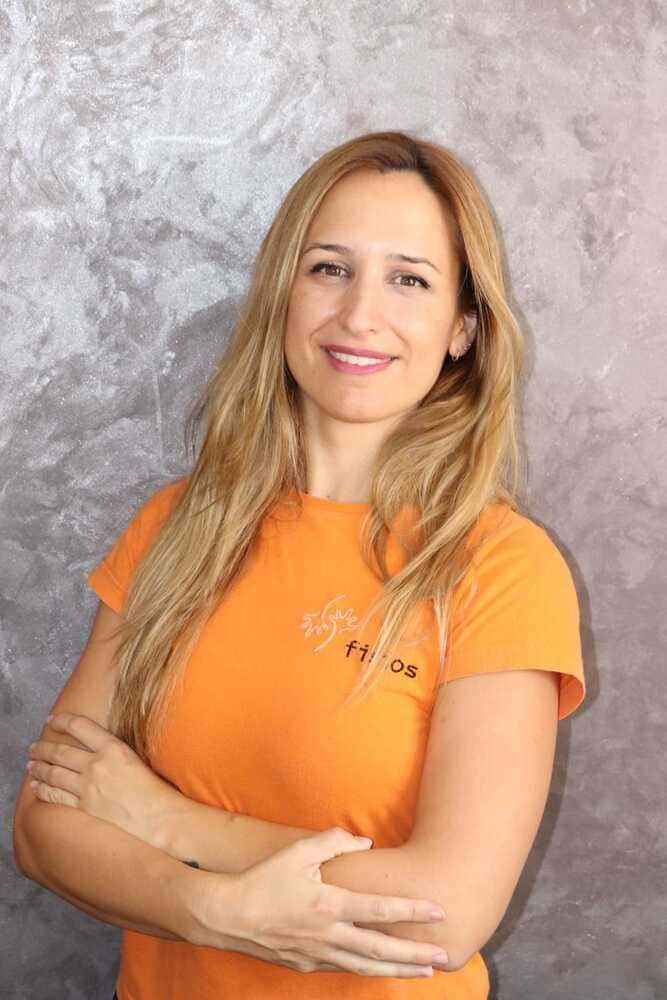 Mujer de cabello rubio, con los brazos cruzados, vistiendo una camisa naranja, sonriendo frente a un fondo gris.