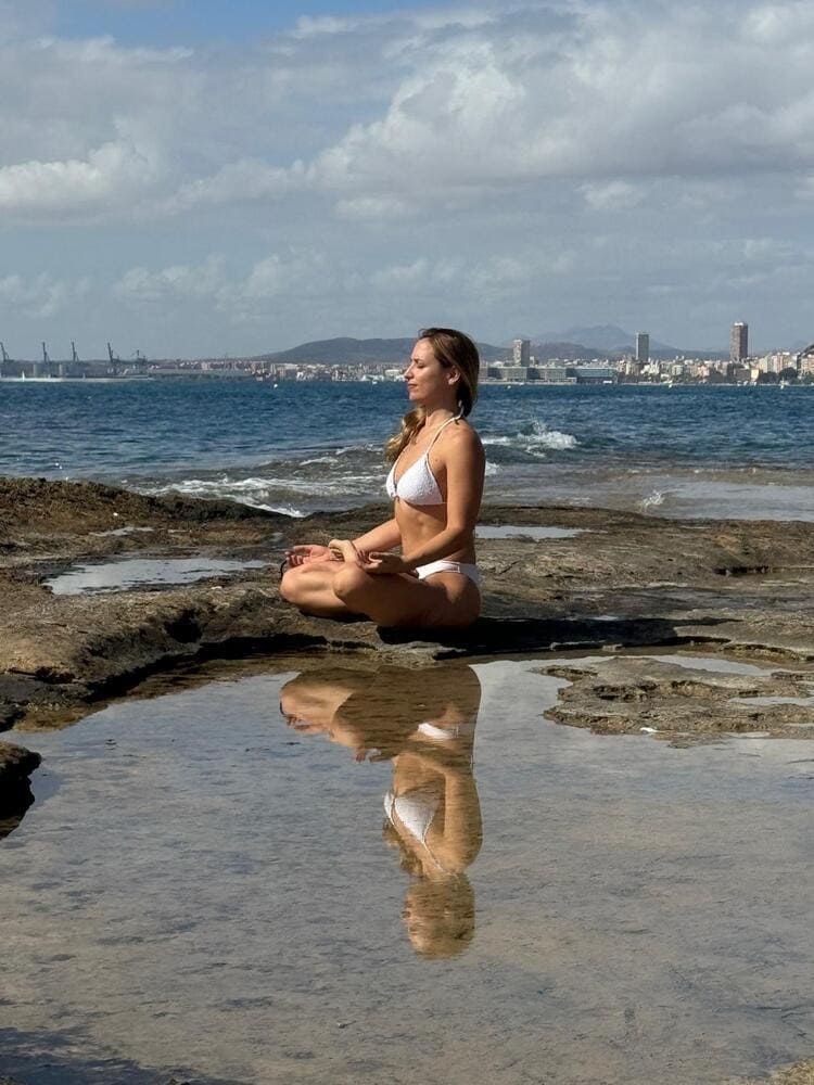 Una mujer en bikini blanco medita en una orilla rocosa, reflejada en un charco. El océano y el horizonte de la ciudad al fondo.