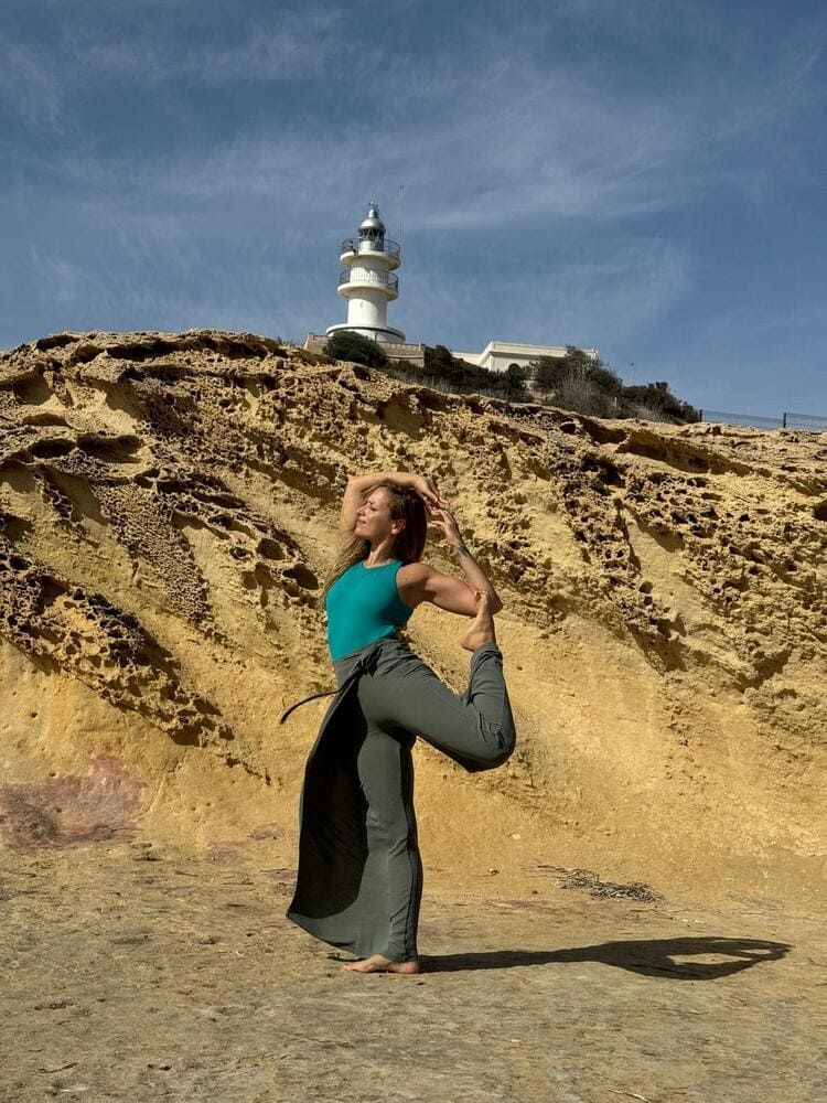 Mujer haciendo yoga junto a un faro en una costa rocosa. Top turquesa, pantalones grises y cielo soleado.