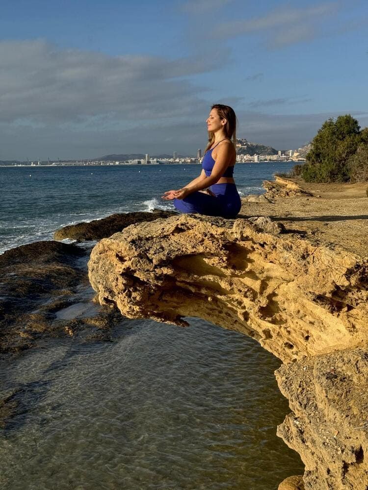 Mujer vestida de yoga azul meditando en un acantilado rocoso con vistas al océano.