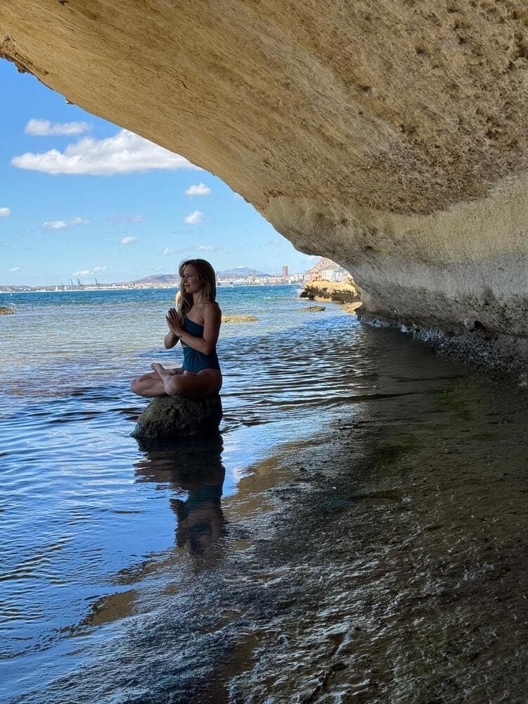 Mujer meditando sobre una roca cerca del borde del agua bajo un acantilado, traje de baño azul, manos en pose de oración.
