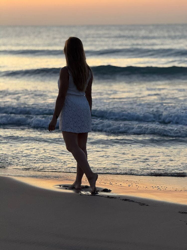 Mujer vestida de blanco camina por una playa al atardecer, con olas al fondo.