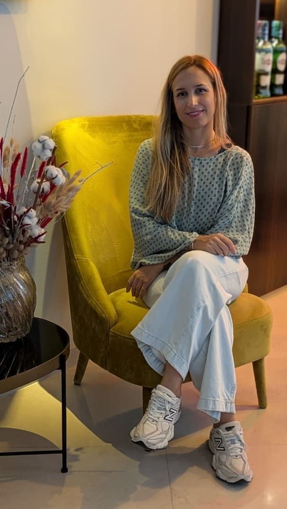 Mujer sentada en una silla amarilla, con blusa verde y jeans claros. Apoyada en un arreglo floral.