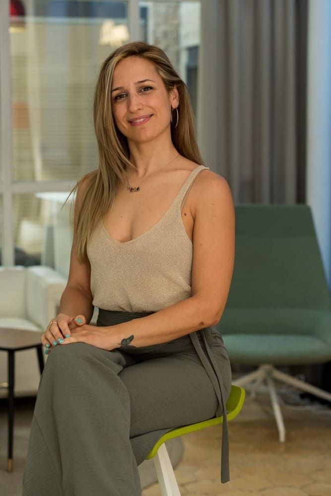 Mujer con cabello largo rubio, con top beige y pantalón verde, sentada, mirando a la cámara.