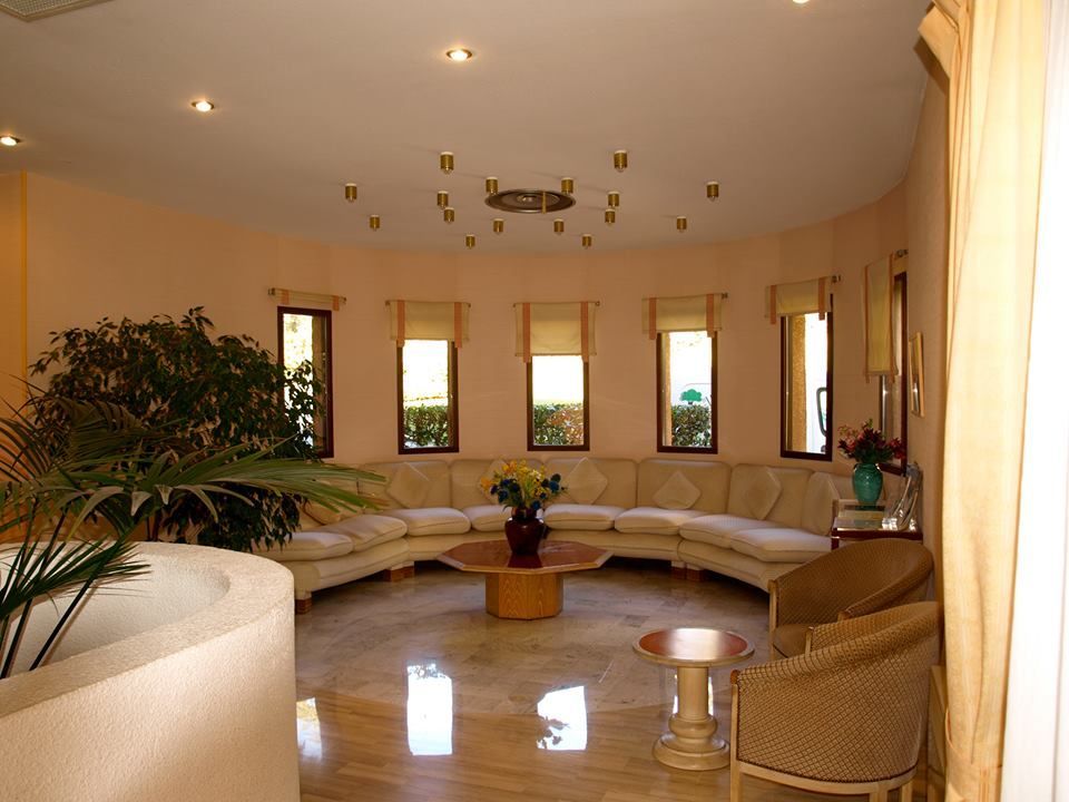 Pièce de forme arrondie avec fenêtres, canapé intégré, table basse et fauteuils. Grande plante et sol en marbre.