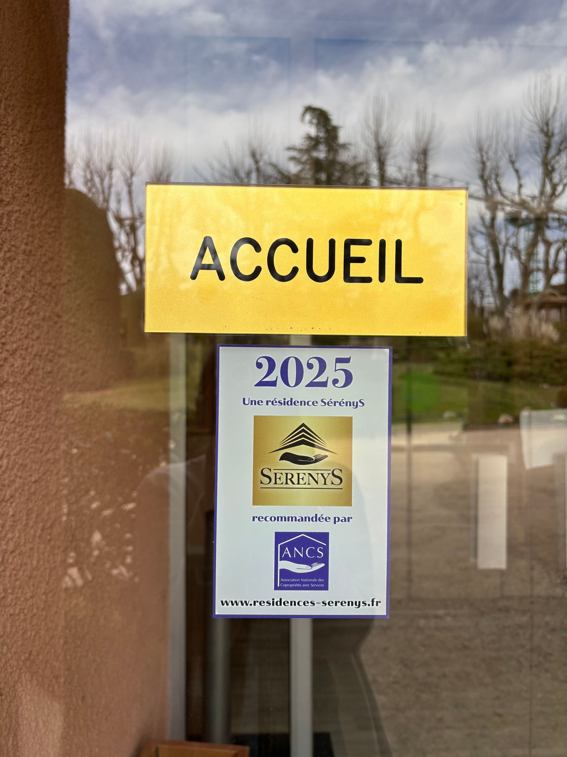 Une porte vitrée ornée d'un panneau jaune ACCUEIL au-dessus d'une affiche des prix Serenys 2025.