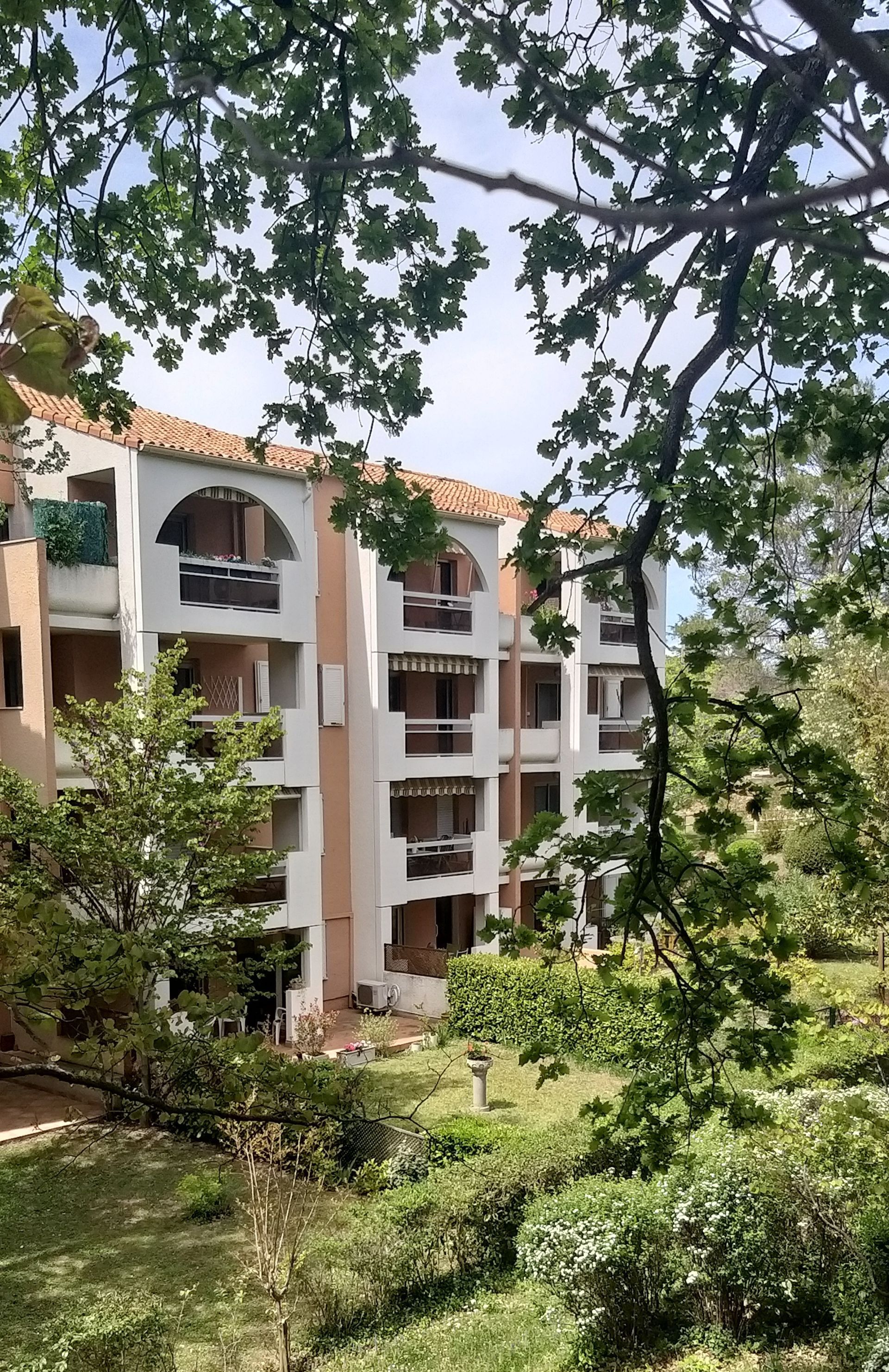 Immeuble d'appartements blanc avec fenêtres cintrées, toit en tuiles de terre cuite, entouré d'arbres verts.