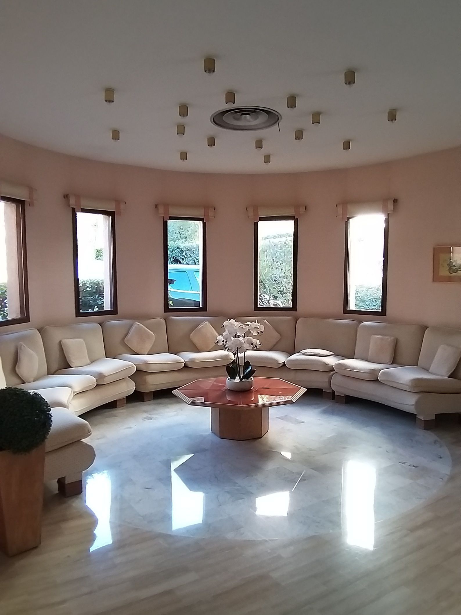 Salon circulaire avec canapés beiges, fenêtres et table centrale.