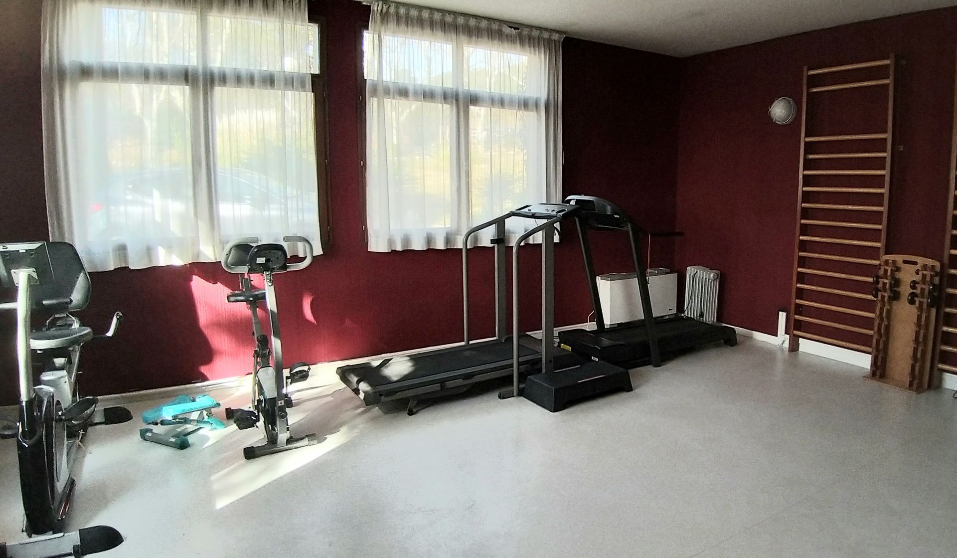 Salle de sport équipée d'appareils de fitness comme un tapis de course et un vélo stationnaire près d'une fenêtre.