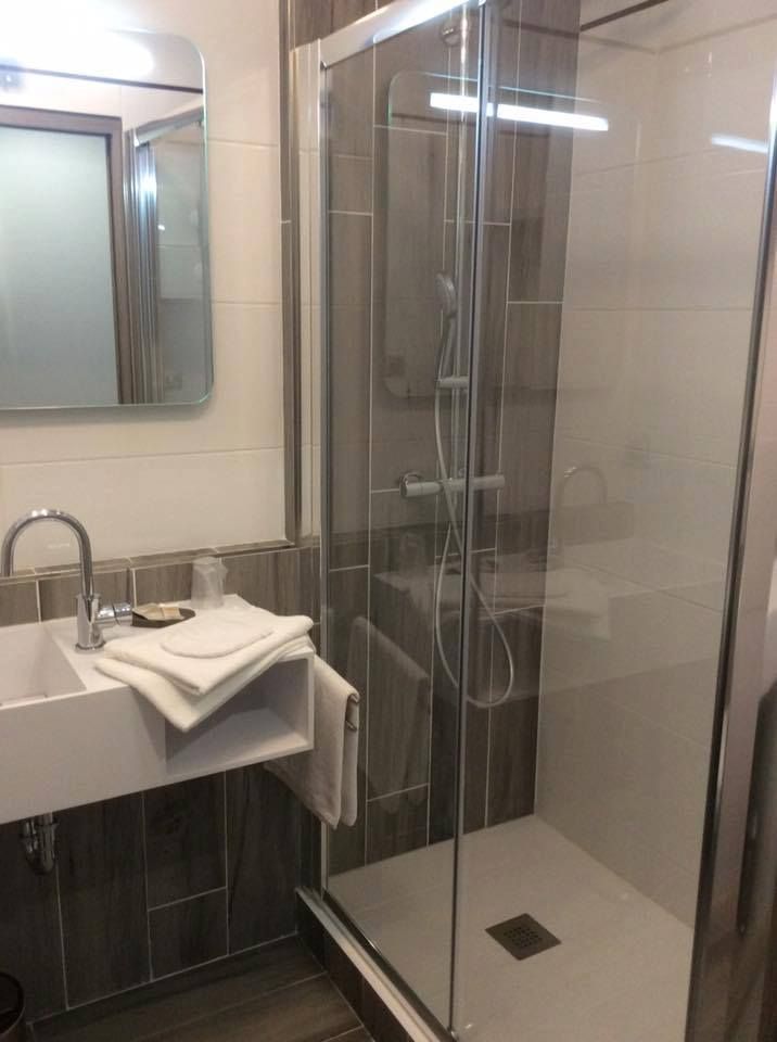 Salle de bains avec douche, lavabo et miroir. Lavabo et serviettes blancs, porte de douche en verre, mur carrelé gris.