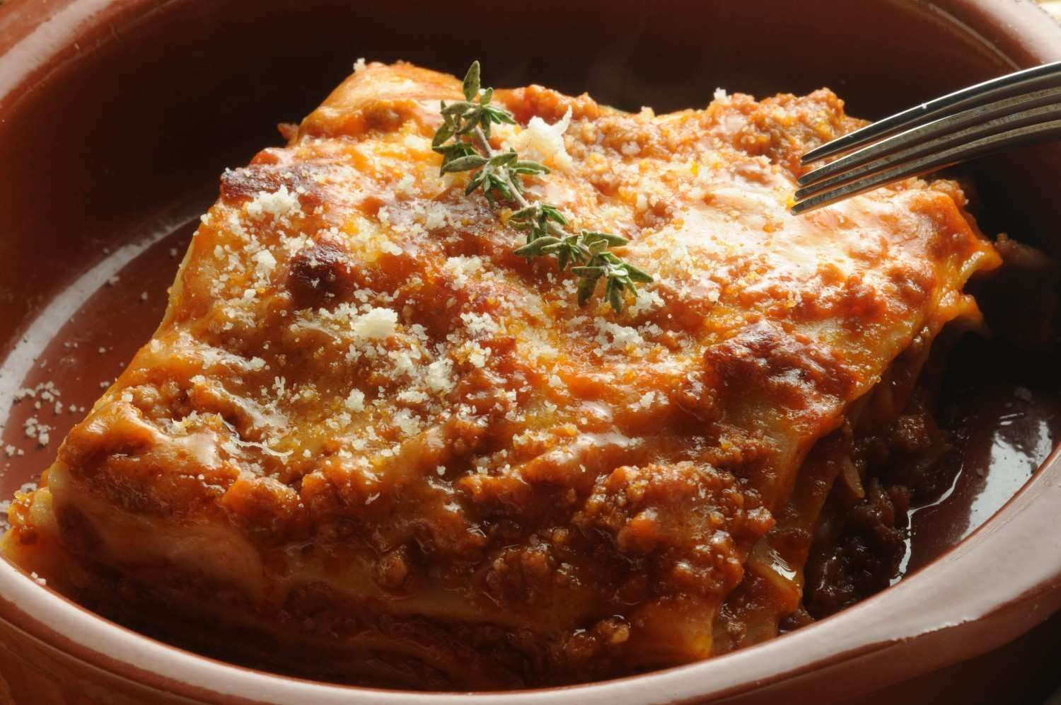 Una porción de lasaña con salsa de carne y parmesano, adornada con una ramita de tomillo en un plato de cerámica.