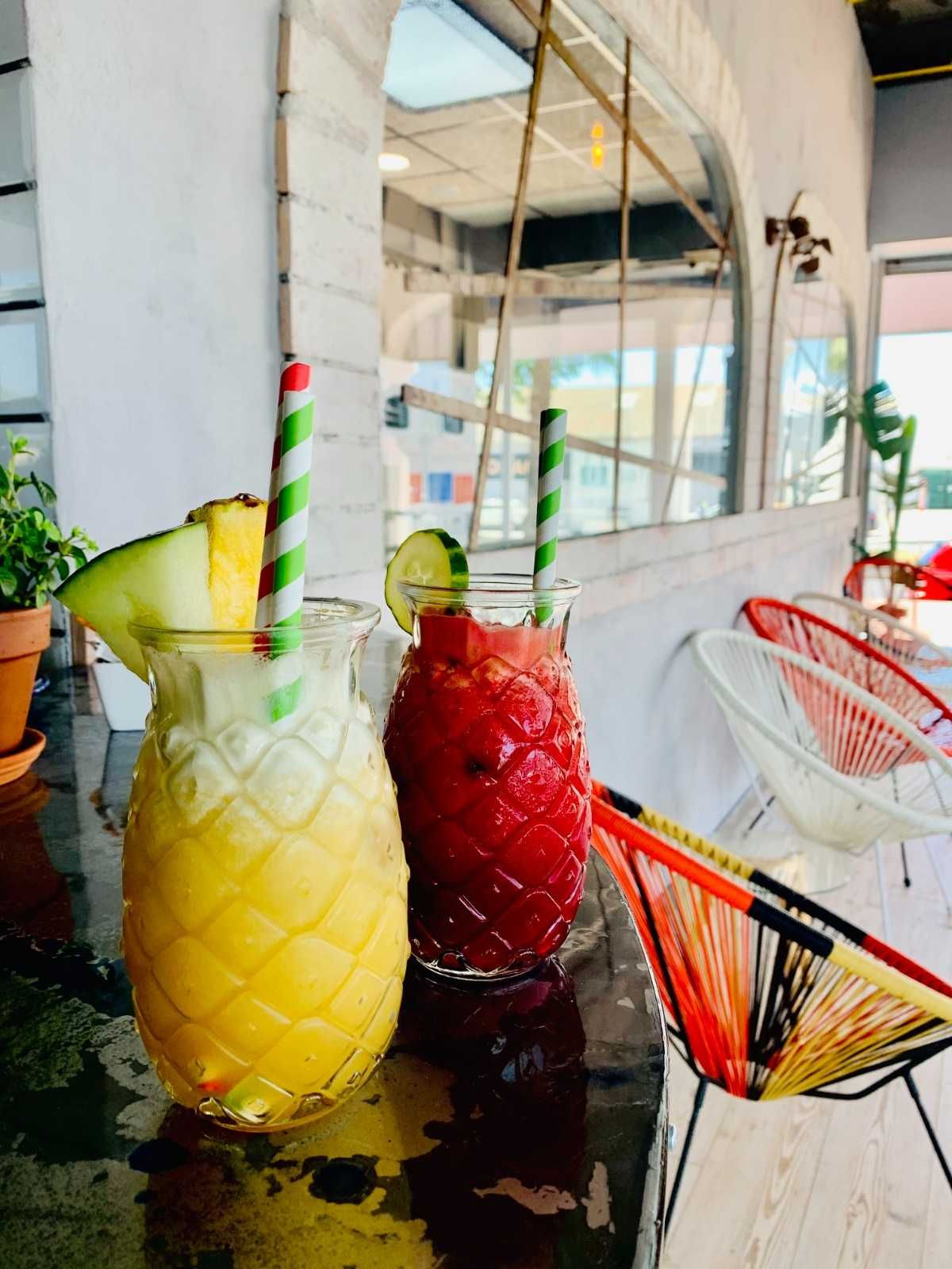 Dos vasos con forma de piña llenos de bebidas de frutas amarillas y rojas, decorados con rodajas de fruta y pajitas a rayas.