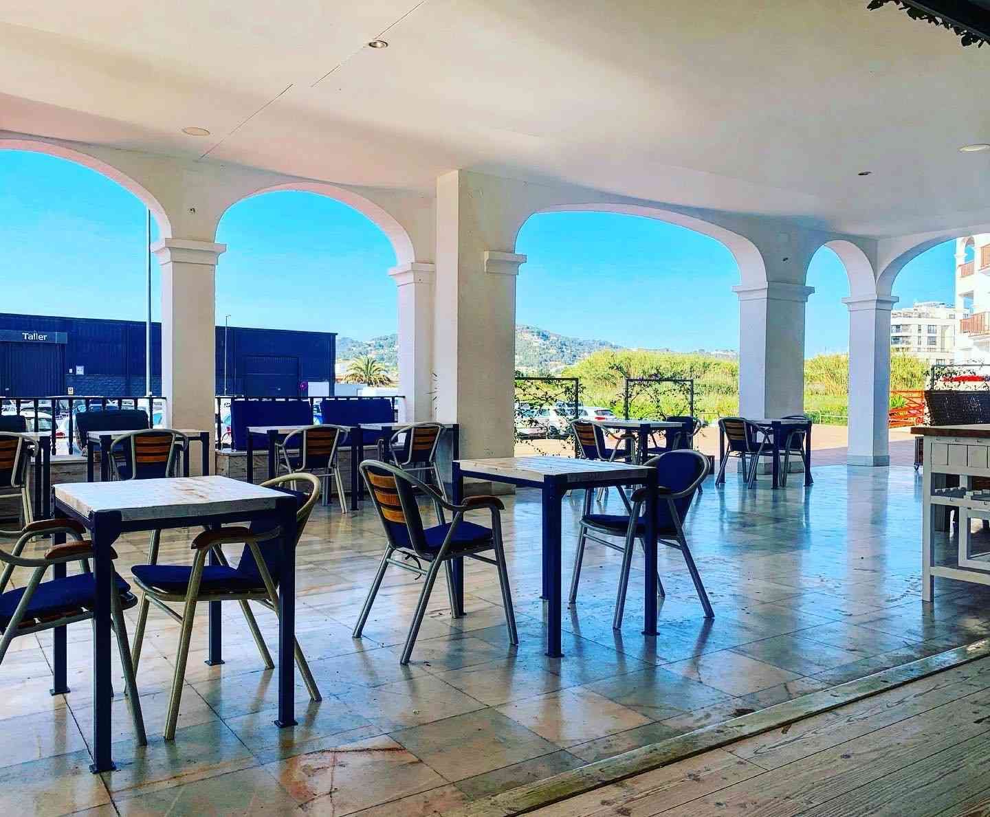 Mesas y sillas vacías en un patio exterior sombreado, con arcos blancos que dan a un paisaje soleado.