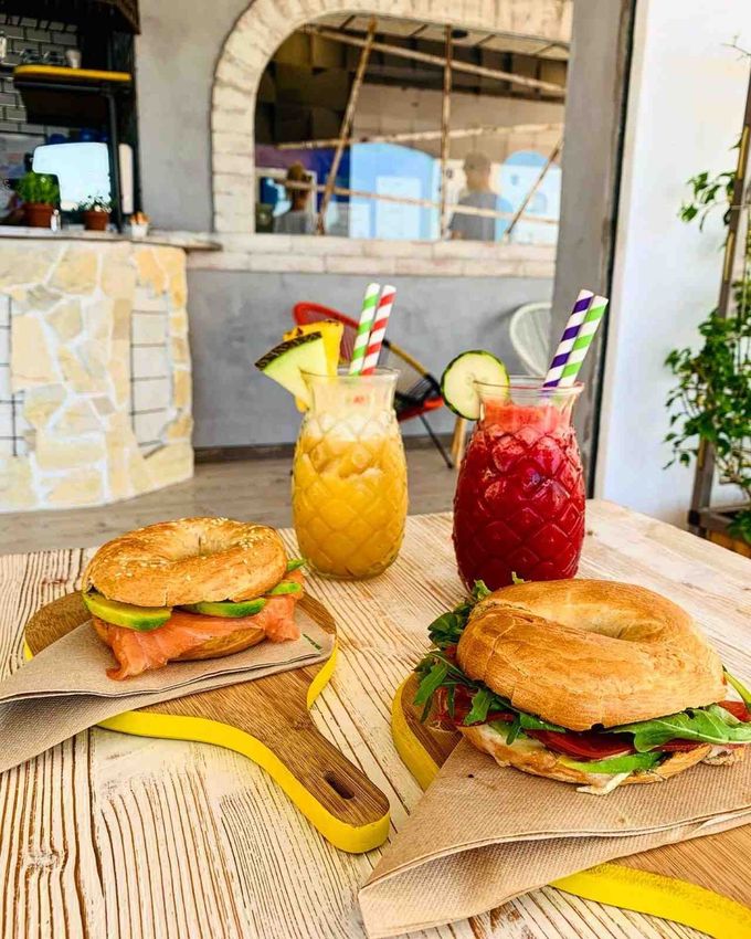 Dos bagels con salmón y verduras reposan junto a un zumo amarillo y un batido rojo en vasos con forma de piña.