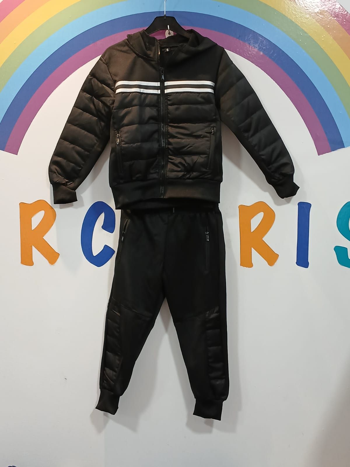 Una chaqueta y unos pantalones negros cuelgan de una percha delante de un arcoíris que dice rcris