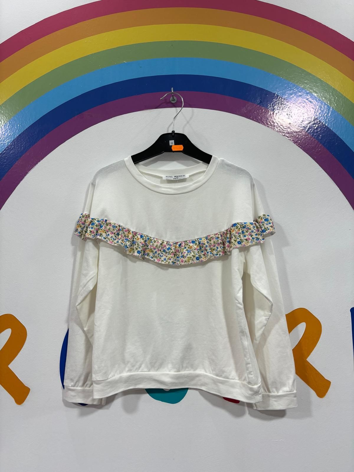 Un suéter blanco con volantes en las mangas cuelga de una percha delante de un arco iris.