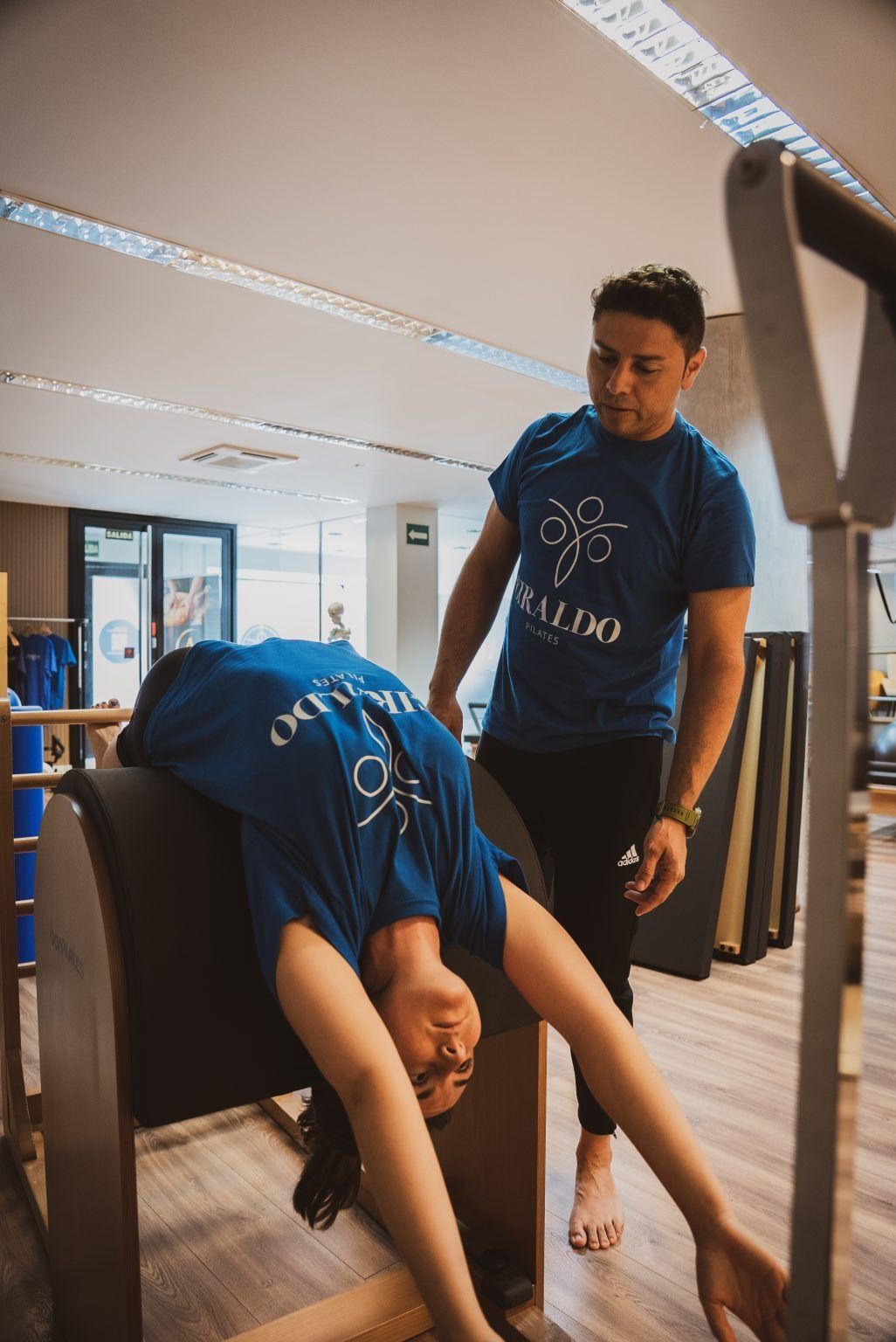 Dos personas se estiran en un gimnasio; una se inclina hacia adelante mientras la otra permanece de pie cerca.