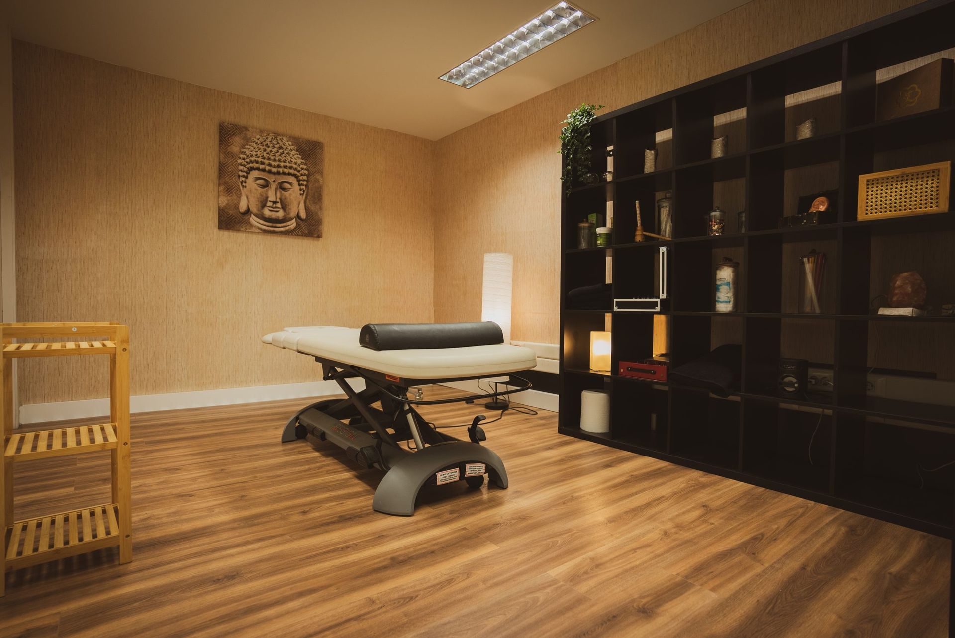 Sala de terapia vacía con una camilla de tratamiento, estanterías negras y suelo de madera bajo una iluminación cálida.