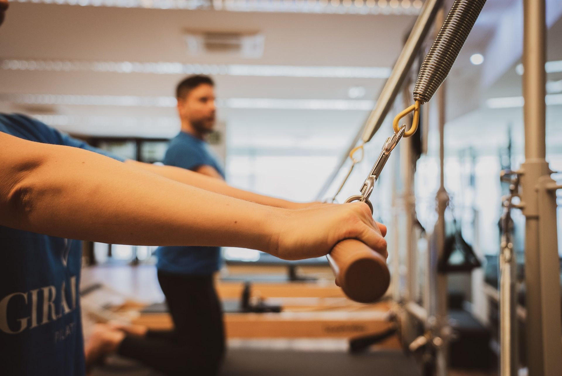 Una persona sujeta la manivela de una máquina de poleas en un gimnasio, con otra persona borrosa al fondo.