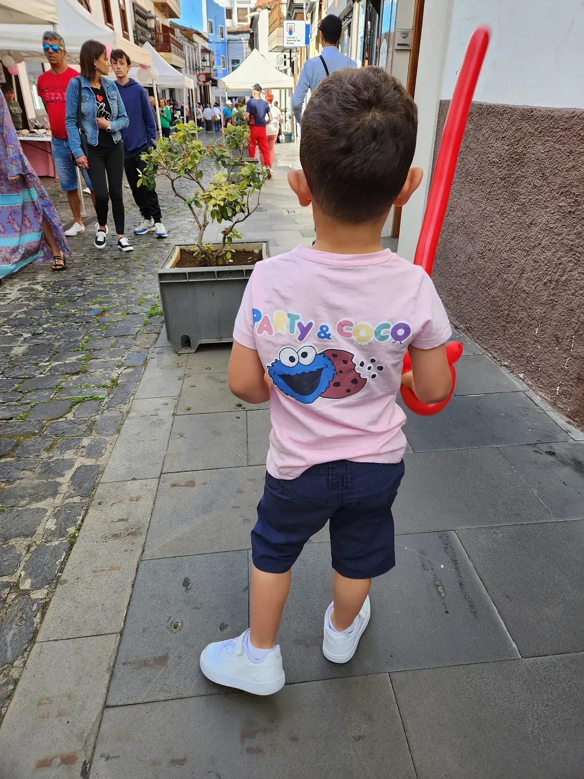 Un niño pequeño que lleva una camiseta de Barrio Sésamo camina por una acera sosteniendo un globo rojo.