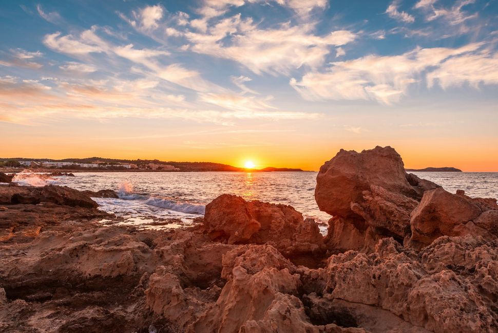 Puesta de sol sobre una costa rocosa con olas y un cálido cielo anaranjado.
