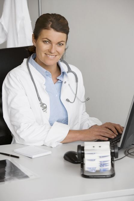 Médica sorrindo em sua mesa, digitando em um laptop, com um estetoscópio, vestindo um jaleco branco.