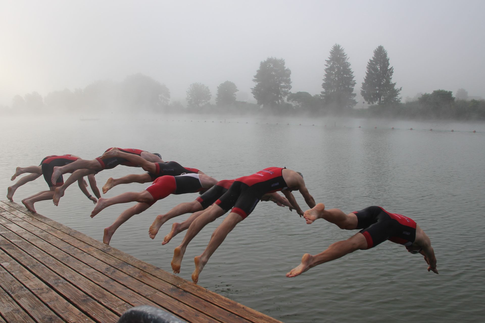 Menschen in Neoprenanzügen springen an einem nebligen Tag von einem Holzsteg ins Wasser.