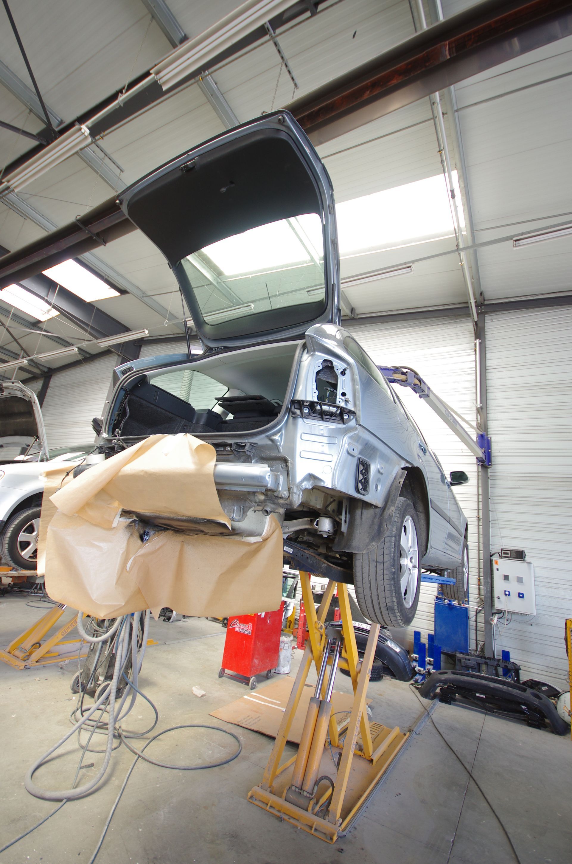 Dans un atelier de réparation, une voiture est soulevée sur un pont élévateur, sa partie arrière de carrosserie ayant été démontée.